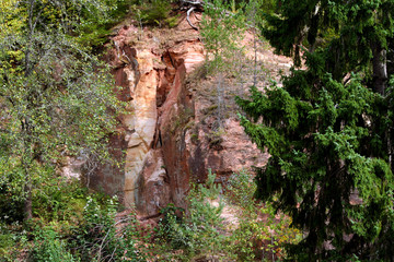 piece of rock red, yellow, brown, white shades, large cliff surrounded by green and yellowing vegetation in autumn, foreground on the left side are tall spruce trees, with the right birch thin trees, 