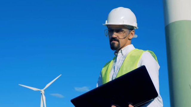 Male energetics worker is standing with his laptop near a wind turbine. Clean, eco friendly energy concept.