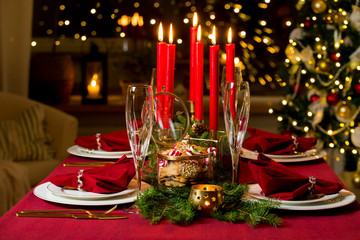 Beautiful served table with candles, Red tablecloth and napkins, white china, gold cutlery, crystal champagne glasses. Living room decorated with lights and Christmas tree. Holiday setting