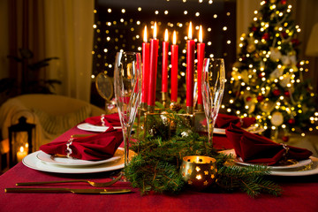 Beautiful served table with candles, Red tablecloth and napkins, white china, gold cutlery, crystal champagne glasses. Living room decorated with lights and Christmas tree. Holiday setting