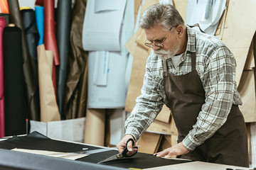mature male handbag craftsman in apron and eyeglasses cutting leather by scissors at workshop