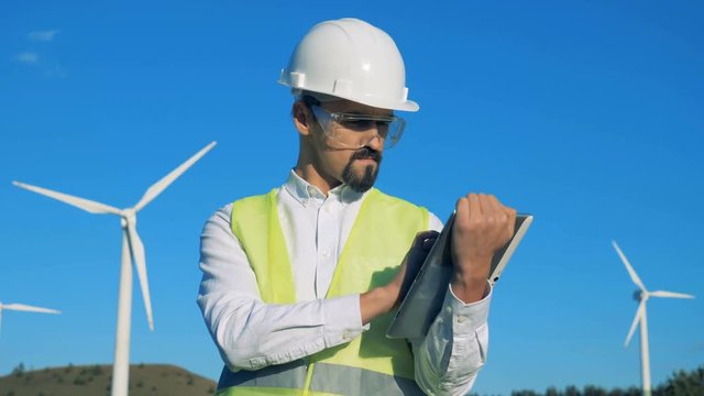 Male worker is operating a tablet while standing beside turbine towers. Clean, eco-friendly energy concept.