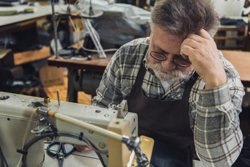 selective focus of mature male tailor in apron and eyeglasses sitting near sewing machine at studio