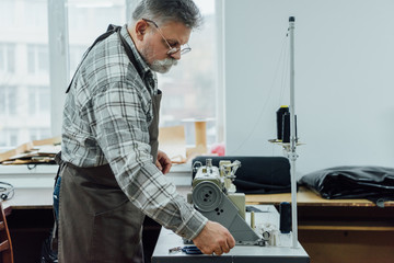 side view of mature male tailor in apron setting strings on sewing machine at workshop