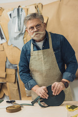 serious middle aged craftsman in eyeglasses and apron looking at camera during work at studio