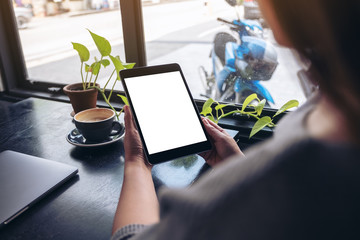 Mockup image of a woman holding and looking at black tablet pc with blank white desktop screen with notebook, green leaves and coffee cup on wooden table