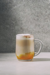 Glass of pumpkin layered spice latte with pumpkin puree, milk foam and cinnamon standing on white marble table with grey wall at background. Copy space