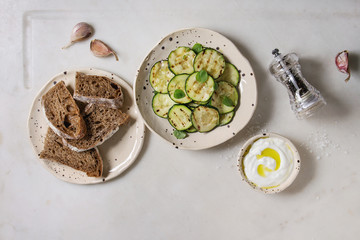 Grilled zucchini salad with yogurt dip and rye sliced bread in spotted ceramic plates over white marble background. Vegetarian food. Flat lay, space
