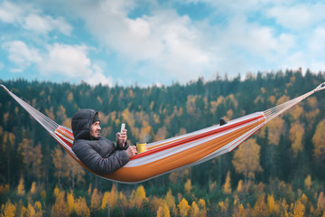 A smiling man sits in a hammock and uses a smartphone in a picturesque location. Mug in his right hand. - obrazy, fototapety, plakaty