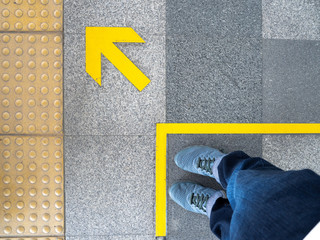 Top view of man feet standing over Arrow symbol on subway platform. Yellow arrow sign on floor at...