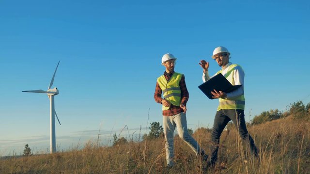 A field with two energetics experts walking across it and a functioning turbine tower. Clean, eco-friendly energy concept.