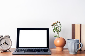 Fototapeta premium Wooden desk with an open laptop mockup, a vase with dried flowers, a concrete gray clock, a mug of coffee and books on the background of a white wall copy space