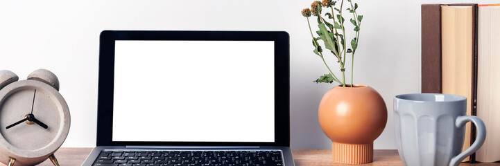 Obraz premium Panoramic photo of wooden desk with an open laptop mockup, a vase with dried flowers, a concrete gray clock, a mug of coffee and books on the background of a white wall