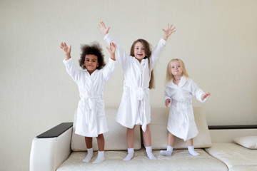 Fototapeta na wymiar Three little girls dancing on the couch. Girls of different nationalities in white coats dance and laugh after a bath.