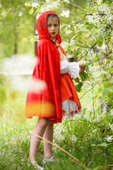 Smiling blond girl posing in a dress of little red riding hood near green trees
