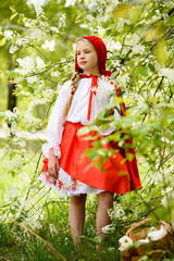 Smiling blond girl posing in a dress of little red riding hood near green trees