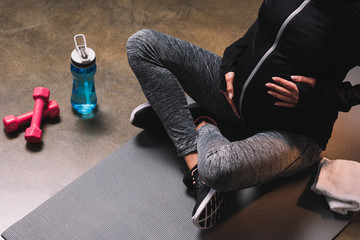 Cropped view of pregnant sporty woman sitting on the sportive mat and sport bottle beside