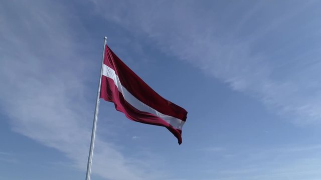 Aerial drone rotating low shot of a large flag of Latvia on blue sky backdrop