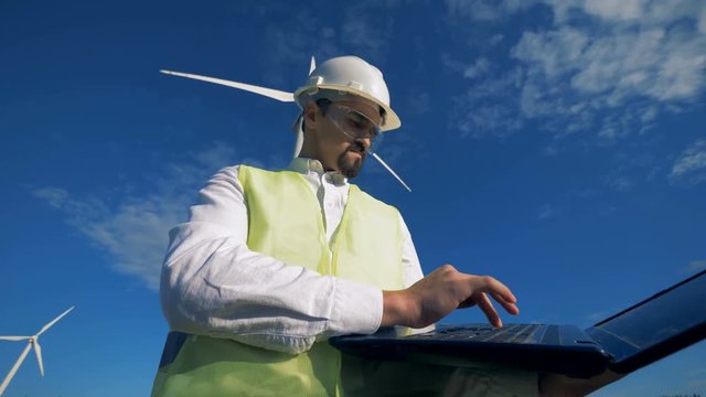 Male specialist is working with a laptop against the background of the sky and windmills