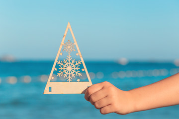 Hand of a child holding a wooden cut out Christmas tree with snowflakes against turquoise sea at the seaside, vacation and travel in winter concept