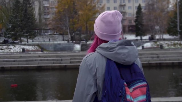 Rear view close-up of hipster woman with backpack in hat. Young beautiful woman with pink hair and pink hat on autumn cityscape background