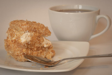 Creamy dessert to treat yourself during a tea break. A piece of a puff pastry cake with a teaspoon on a white plate and a white cup in the background, close up view. Toned.