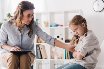 adult psychologist with clipboard talking to sad little child in office