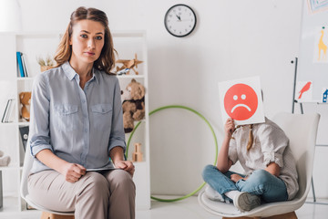 psychologist sitting near little child while she covering face with sad emotion face card
