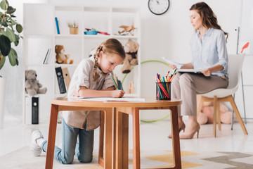 little child drawing while psychologist with clipboard sitting blurred on background