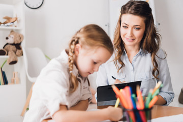 adult psychologist writing in clipboard while looking at little drawing child