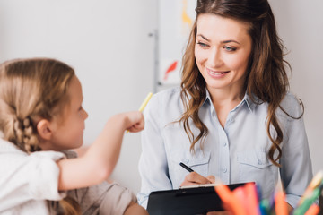 happy psychologist with clipboard sitting near little child and chatting while she drawing with color pencils