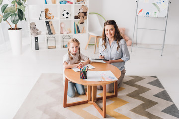 Obraz premium happy psychologist with clipboard sitting near little child while she drawing with color pencils and looking at camera