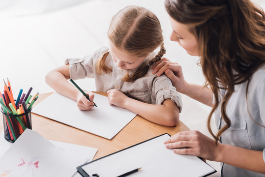 high angle view of psychologist with clipboard sitting near little child while she drawing with color pencils