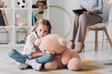 sad little child with teddy bear sitting on floor with psychologist sitting on background