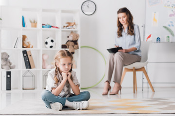 depressed little child sitting on floor with psychologist sitting on background