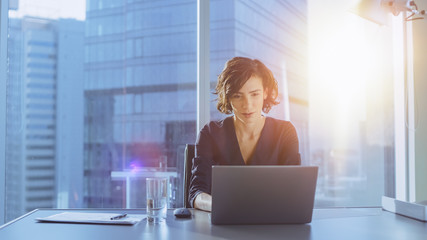 Beautiful Successful Businesswoman Working on a Laptop in Her Office with Cityscape View Window. Strong Independend Female CEO Runs Business Company. Sun Flares Behind Her. - obrazy, fototapety, plakaty