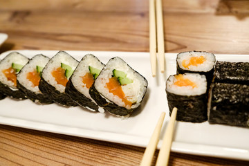 Horizontal shot of a close-up of rice rolls with salmon, cucumber and cream cheese on a white porcelain stretched rectangular dish on a wooden table in natural light colors.