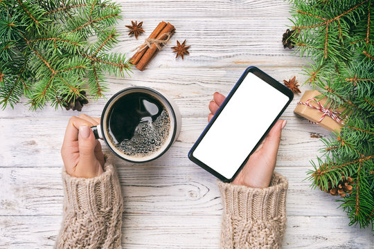 Female hands holding modern smartphone with mosk up and mug of coffee on wooden rustic vintage table with christmas decoration. top view