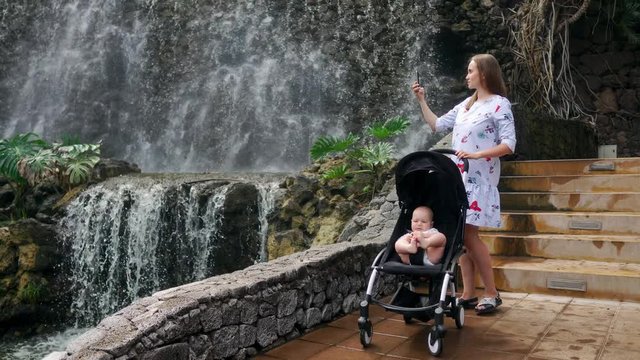 A young mother walking with her baby near the waterfall takes pictures on her smartphone while traveling with her family.