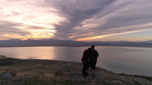Silhouette of couple standing on mountain top at sunset revealing them while fling by as they cuddle.