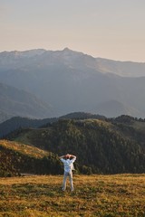 A man is standing on the peak of a cliff with his back to the camera, enjoying a scenic view of the green forest and beautiful mountains.