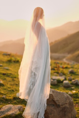 Fototapeta na wymiar fine-art wedding. Beautiful girl with gorgeous slim in gray dress with hand-painted flowers stands against the mountain landscape at sunset