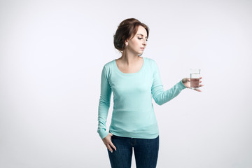 Beautiful girl with brown-haired woman dressed in jeans and mint-colored sweater on a light background holds in her left hand a glass of pure drinking water  and Looks at the glass.. 
