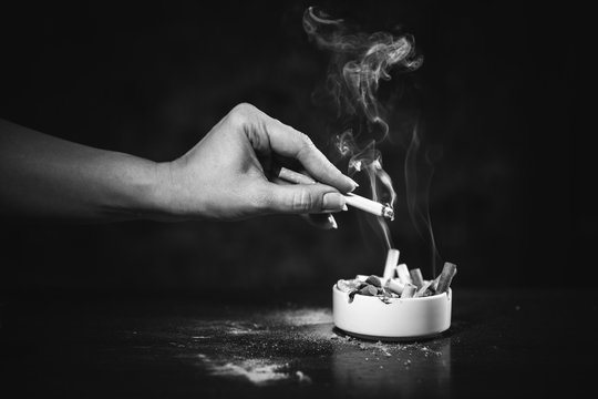 Black white photo. A woman's hand shakes off the ashes of a burning cigarette in an ashtray, where there are a lot of cigarette butts. Smoking is dangerous for women's health. Cigarette Smoke.