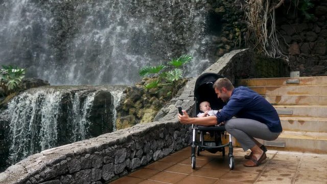 A young father walking with a child in a wheelchair near the waterfall takes pictures on his smartphone for social networks and posting on the Internet while traveling with his family. The concept of