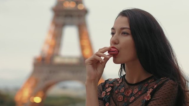 Paris woman smiling eating the french pastry macaron in Paris. Evening Eiffel tower with lights in the background. Portrait of gorgeous romantic young sensual girl in fashion black sexy dress enjoying