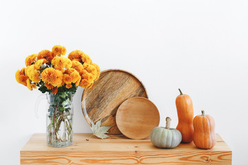 Different multicolored decorative pumpkins, wooden plates or dishes and vase with yellow chrysanthemums flowers on a wooden table on a background of white wall. Autumn or fall home interior decor.