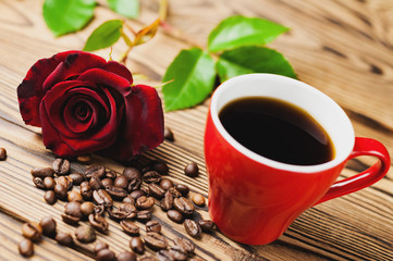 Full cup of black coffee near one red rose and scattered coffee beans on old wooden table. Saint Valentine's Day concept