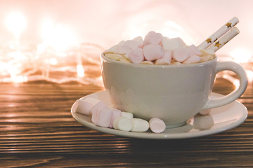hot chocolate with marshmallow candy on wooden background light from garland, close-up selective focus