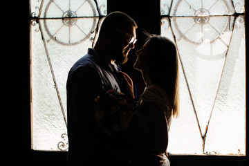 portrait of a romantic couple in a backlight from a window or door, silhouette of a couple in a doorway with a backlight, couple of lovers groom and bride at the window, girl holding flowers in hands